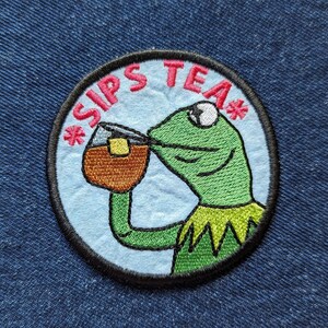 May include: A circular embroidered patch featuring a green frog character drinking from a brown mug. The background is light blue with a black border. The text *SIPS TEA* is embroidered in pink letters along the top curve of the circular patch.