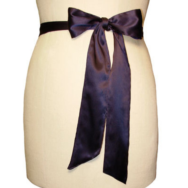 May include: A dark purple satin ribbon tied in a bow around a waist. The ribbon is long and flowing, with the ends trailing down the sides.