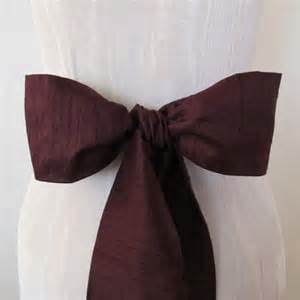 May include: A burgundy satin sash with a bow tied in the center. The sash is tied around a white dress.