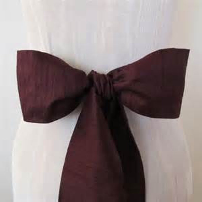 May include: A burgundy satin sash with a bow tied in the center. The sash is tied around a white dress.