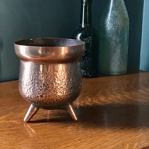 May include: A hammered copper planter with three legs. The planter is on a wooden surface with two glass bottles behind it.