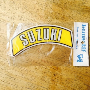 May include: A yellow sew-on patch with the word "SUZUKI" in white letters. The patch is packaged in a clear plastic bag with a white label that says "Leomotïf Sew-on Patches".