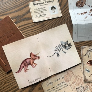 May include: A paleontologist field guide with a brown leather cover. The guide features a watercolor illustration of a Triceratops dinosaur with a skeleton illustration of the same dinosaur on the opposite page. The text "Triceratops" is printed below the illustration. The guide also includes a page with the text "Cody turns 6. Come have a ROARING good time and help dig dinosaur bones!"