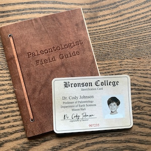 May include: A brown leather-bound journal with the title "Paleontologist Field Guide" embossed on the cover.  A white card with the text "Bronson College Identification Card" and a photo of a young boy is next to the journal.