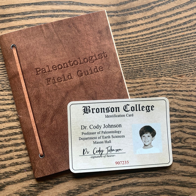 May include: A brown leather-bound journal with the title "Paleontologist Field Guide" embossed on the cover.  A white card with the text "Bronson College Identification Card" and a photo of a young boy is next to the journal.