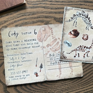 May include: A tan and brown invitation card with a dinosaur skeleton illustration. The text on the card reads "Cody turns 6. Come have a ROARING good time and help dig up some dinosaur bones! Excavation Date/Time: July 8, 2-4 pm. Excavation Location: Cody's House. RSVP: 555-657-0987. Come ready to get a little dirty!"
