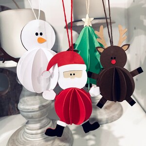 May include: Three paper ornaments hanging from a string. A white snowman ornament, a red Santa Claus ornament, and a brown reindeer ornament. The ornaments are all 3D and have a layered paper design.
