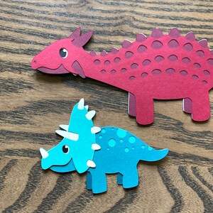 May include: Two paper cut-out dinosaurs. A red dinosaur with a spiky back and a blue dinosaur with three horns on its head.