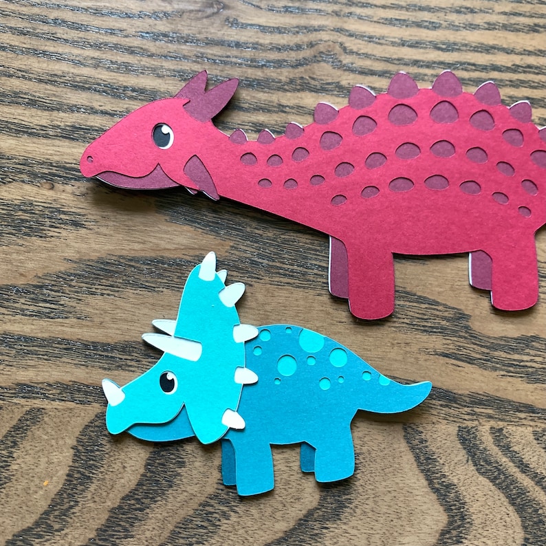 May include: Two paper cut-out dinosaurs. A red dinosaur with a spiky back and a blue dinosaur with three horns on its head.