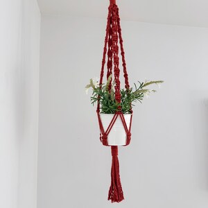 May include: A red macrame plant hanger with a white ceramic pot filled with greenery and white flowers. The hanger is suspended from a wooden ring and features a long tassel. The plant holder is hanging against a white wall.