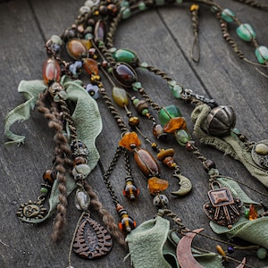 Könnte beinhalten: Nahaufnahme einer Sammlung von Schmuck im Bohème-Stil. Die Stücke weisen eine Vielzahl von Perlen in Bernstein-, Grün- und Brauntönen sowie Metallanhänger auf. Der Schmuck ist auf einem Holztablett mit grünen Stoffakzenten angeordnet.