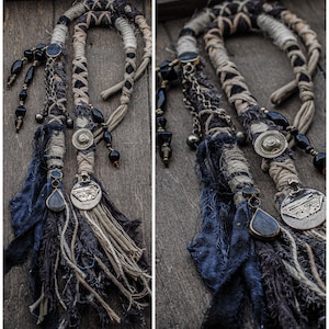 Peut inclure: Un accessoire de coiffure composé de cordons en tissu beige et noir tressés et entourés de ficelle. Il comprend des perles noires pendantes, des chaînes métalliques, des breloques circulaires argentées et des bandes de tissu effilochées bleu foncé.