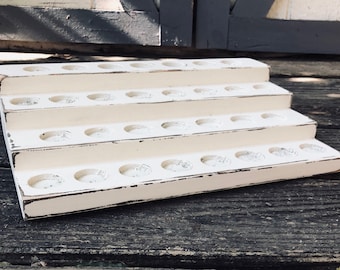 May include: A tiered, off-white wooden display stand with distressed edges. Each tier has multiple circular cutouts. The stand is designed to hold small items and is set against a rustic wooden background.