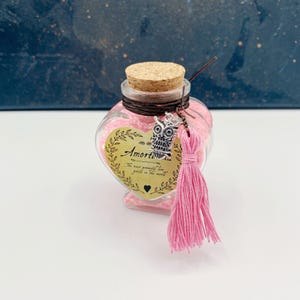 May include: A heart-shaped glass bottle filled with pink glitter and a cork stopper. The bottle has a brown twine tied around the neck and a silver owl charm with a pink tassel hanging from it. The bottle has a label that reads "Amortentia The most powerful love potion in the world".