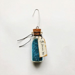 May include: A small glass bottle with a cork stopper filled with blue glitter. The bottle has a silver wire loop for hanging and a tag that reads "Drink Me".