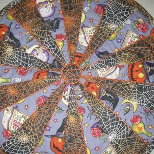 May include: A circular quilted fabric mat featuring a pieced design with alternating wedges. One set of wedges displays a light purple background with printed pumpkins, potion bottles, roses, and books. The other wedges feature black spiderwebs.