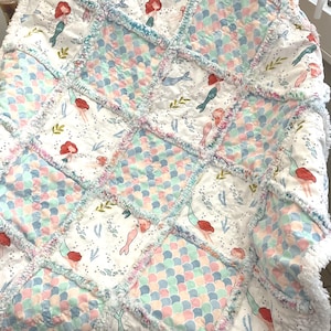 May include: A handmade, patchwork quilt featuring a mermaid theme. The quilt showcases a white background with colorful mermaid, jellyfish, and seaweed patterns. The squares are connected with a ruffled, fringed edge, and the quilt is soft and inviting.
