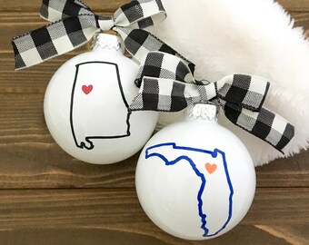 May include: Two white Christmas baubles with black and white buffalo check ribbon. One bauble features a black outline of Alabama with a red heart in the centre. The other bauble features a blue outline of Florida with an orange heart in the centre.