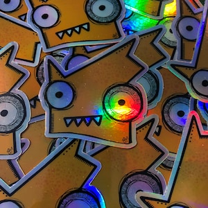 Puede incluir: Un montón de pegatinas holográficas con la cara de un monstruo de dibujos animados con un ojo grande, dientes de sierra y forma de corona. Las pegatinas tienen un fondo amarillo-naranja y un acabado reflectante con efecto arcoíris.