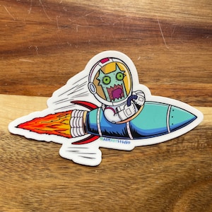 Puede incluir: Una pegatina con un astronauta alienígena de dibujos animados en un cohete. El alienígena tiene piel verde, ojos grandes y la boca abierta. El cohete es azul con una llama roja y naranja. La pegatina tiene el texto "ABMARTstudio".