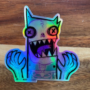 May include: Holographic sticker featuring a cartoon monster with button eyes and a skeletal body. The monster has a wide, toothy grin and outstretched hands. The sticker has a rainbow sheen and the artist's signature, ABMArtStudio, is visible.