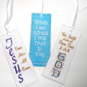 May include: Three white fabric bookmarks with embroidered text. The first bookmark says "Jesus" in purple lettering and "Name Above All Names" in gold lettering. The second bookmark is light blue and says "When I am Afraid I Will Trust In You" in gold lettering. The third bookmark says "Be Still and Know That I AM GOD" in gold lettering.