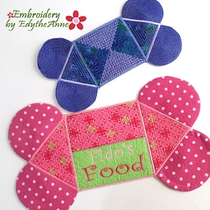 Könnte beinhalten: Zwei Stoffboxen in Form eines Hundebeins, eine blaue mit geometrischem Muster und eine rosa mit weißen Punkten. Die rosa Box hat den Text "Fido's Food" auf der Vorderseite gestickt.