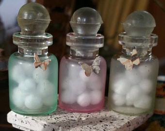 Puede incluir: Tres botellas de vidrio con tapones de vidrio transparente y objetos redondos blancos en su interior. Las botellas están llenas de objetos redondos blancos y tienen un acabado esmerilado. Las botellas están dispuestas sobre una superficie blanca texturizada. Las botellas tienen un acabado de vidrio azul claro, rosa y transparente.