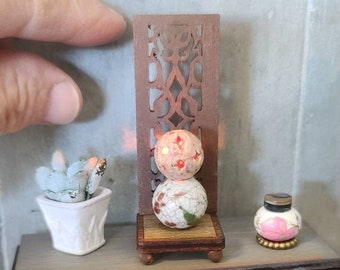 以下が含まれることがあります： 正面に時計の文字盤のデザインが施されたミニチュアの木製テーブル。テーブルには、小さな鉢植えの多肉植物、白と金の球体を持つ装飾的なランプ、そして小さなピンクと金のボトルが置かれています。