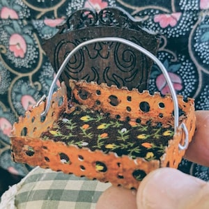 Puede incluir: Un marco de cama de metal naranja en miniatura con un colchón estampado floral y un asa de alambre. El marco de la cama tiene un diseño decorativo recortado.