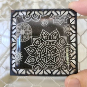 May include: Square black decorative tile with a detailed white mandala design. The tile is held by a hand and has a decorative black frame. The mandala design includes a central star and various floral and sun-like patterns.
