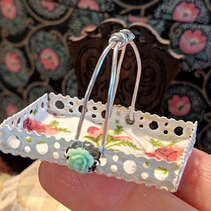 Puede incluir: Una cesta de metal blanco con un forro de tela floral y un adorno floral verde en el asa. La cesta es pequeña y parece estar hecha a mano.