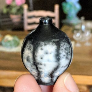 以下が含まれることがあります： 小さくて丸い、白と黒の装飾的な花瓶。 花瓶は暗く光沢のある上部と、白とグレーの斑点模様があります。 花瓶は人の手で持たれています。