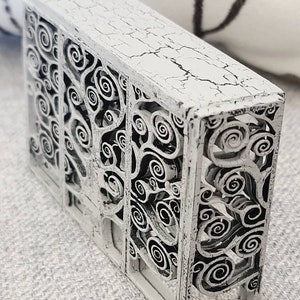May include: A white wooden box with a decorative design of swirling branches and leaves. The box has a distressed, aged look with a white paint finish.