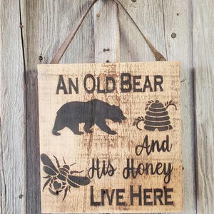 May include: A rustic wooden sign with the text "An Old Bear And His Honey Live Here". The sign features a black silhouette of a bear, a honeycomb, and a bee.