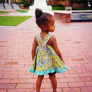 May include: A young girl wearing a green and blue floral dress with a ruffled hem stands on a brick walkway. She is looking to the right of the frame. The background features a fountain and a brick building.