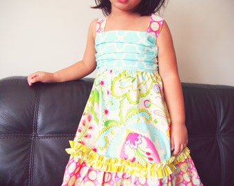 May include: A young girl wearing a colorful, floral print dress with a blue and white patterned bodice. The dress has a ruffled hem and is perfect for a summer party.