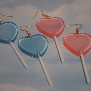 May include: Three pairs of heart-shaped lollipop earrings. One pair is pink, and two pairs are blue.