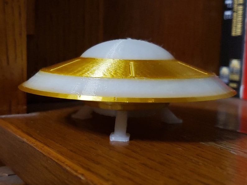 K&ouml;nnte beinhalten: Ein wei&szlig;es und goldenes Modell einer fliegenden Untertasse mit drei Beinen. Die Untertasse hat einen goldenen Rand und eine wei&szlig;e Unterseite.