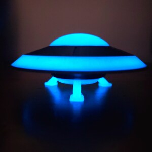 K&ouml;nnte beinhalten: Ein blaues und schwarzes UFO-f&ouml;rmiges Nachtlicht mit drei Beinen. Das Licht ist an und leuchtet blau.