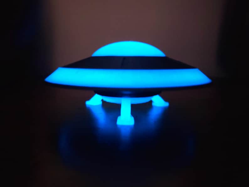 K&ouml;nnte beinhalten: Ein blaues und schwarzes UFO-f&ouml;rmiges Nachtlicht mit drei Beinen. Das Licht ist an und leuchtet blau.