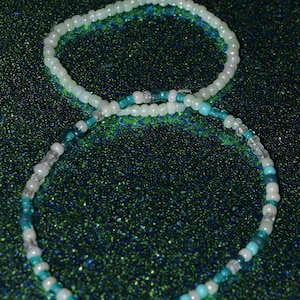 May include: Two beaded necklaces, one white and one with turquoise and white beads, on a green sparkly background.