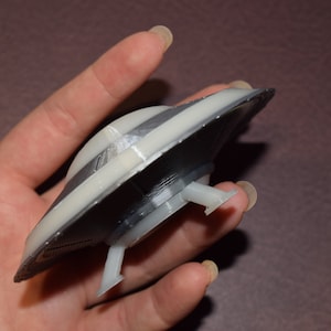 K&ouml;nnte beinhalten: Ein 3D-gedrucktes Modell einer fliegenden Untertasse mit einem Grau- und Wei&szlig;farbschema. Die Untertasse hat drei Landebeine.