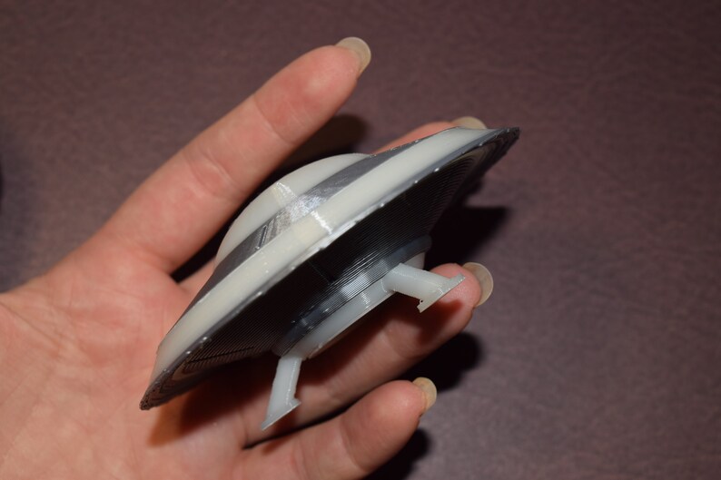 K&ouml;nnte beinhalten: Ein 3D-gedrucktes Modell einer fliegenden Untertasse mit einem Grau- und Wei&szlig;farbschema. Die Untertasse hat drei Landebeine.