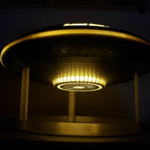 May include: A small, silver-colored UFO-shaped lamp with a white base and a ring of yellow lights around the bottom of the saucer.