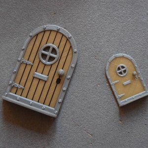 May include: Two miniature fairy doors. The doors have a light brown wooden appearance with vertical black lines, a circular window, and a silver frame. One door is larger than the other.