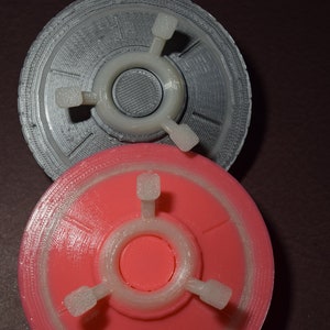 K&ouml;nnte beinhalten: Zwei 3D-gedruckte Objekte in Form einer fliegenden Untertasse. Das obere Objekt ist silberfarben und das untere Objekt ist rosa.