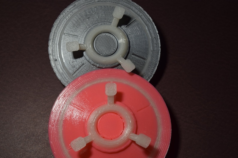 K&ouml;nnte beinhalten: Zwei 3D-gedruckte Objekte in Form einer fliegenden Untertasse. Das obere Objekt ist silberfarben und das untere Objekt ist rosa.