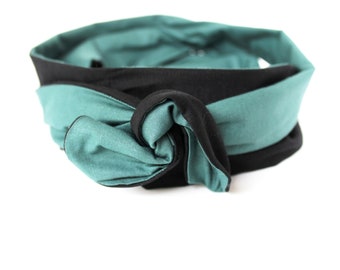 May include: A reversible headband featuring a teal and black color scheme. The headband is knotted at the front, showcasing the contrasting colors. The teal fabric is visible on the outside, while the black fabric lines the inside.