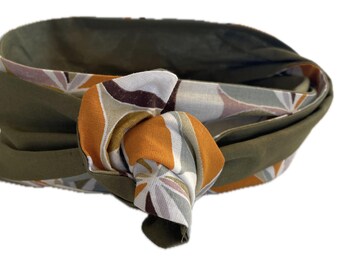 May include: Olive green fabric headband with a knotted design. The knot features a patterned fabric with orange, brown, and white geometric shapes. The headband is designed to be worn around the head.